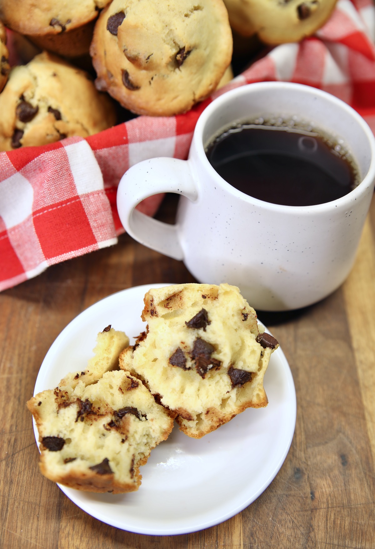 Chocolate chip muffin on a plate, split open, cup of coffee, basket of muffins.