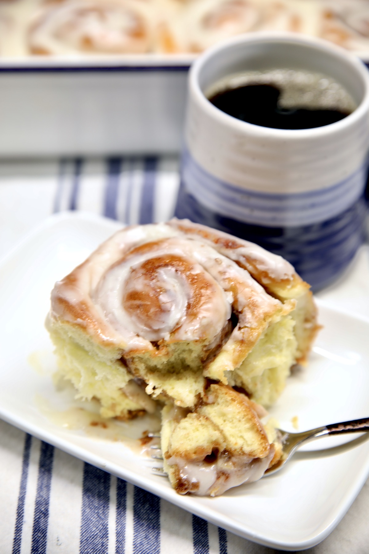 Cinnamon roll on a plate with bite on a fork, cup of coffee.