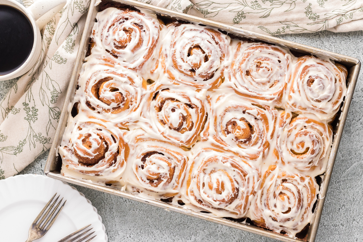 Frosted cinnamon rolls in a pan with plates and linens. 